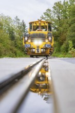 Yellow machine on rails with reflection in the foreground, surrounded by trees, tamping machine,