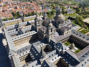 Aerial view of a historic monastery with towers and nearby town, surrounded by green landscape,