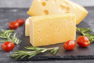 Various types of cheese with rosemary and tomatoes on black slate board on a black concrete