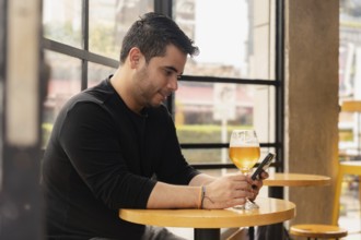 Profile of Latin man sitting enjoying a glass of beer while texting with his smartphone in a pub