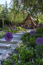 View of a garden with a wooden hut and a stone path, lined with purple flowers and surrounded by