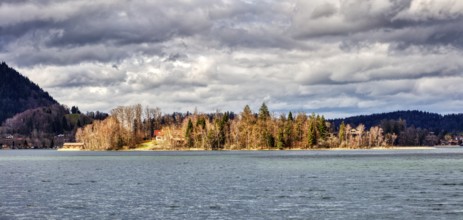 Island with trees in autumn light, surrounded by a calm lake under a cloudy sky, Schliersee