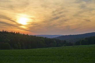 Sunset over a green meadow, surrounded by forest and hills under a cloudy sky, summer, Mönchberg,