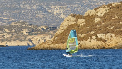 Person windsurfing along a rocky coast on blue water, windsurfer, Meltemi windsurfing spot, Devils