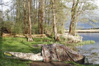 A canoe with camouflage net on the lakeshore, surrounded by trees and quiet, natural surroundings,