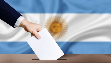 Hand holding ballot in voting ballot box with Argentina flag in background. Hand man puts ballot