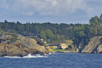 Coastal view with a wooded hill, rocks on the shore, several houses and waves under a cloudy sky,