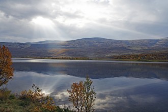 A lake with mountains in the background, rays of light penetrate the clouds, autumn leaves and