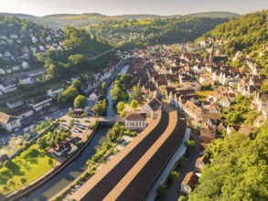 Aerial view of a town with many roofs and a river with a bridge, surrounded by green hills in