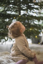 A small child sits in front of a decorated Christmas tree with lights in a cosy room and looks up