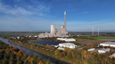 Demolition of a chimney with smoke cloud, surrounded by autumn landscape and wind turbines,