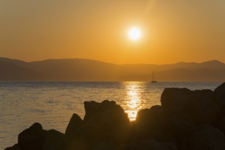 Sunset over the sea with a sailing boat in the distance, rocks in the foreground, Agíos Nikólaos,