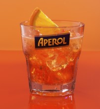 Aperol on ice cube cubes with half an orange slice in an aperitif tumbler against an orange