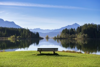 A calm lake surrounded by mountains, with a bench in the foreground and a clear reflection in the