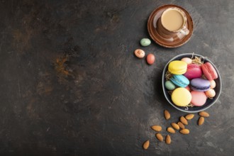 Multicolored macaroons and chocolate eggs in ceramic bowl, cup of coffee on black concrete