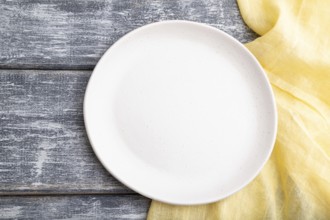 Empty white ceramic plate on gray wooden background and yellow linen textile. Top view, close up,