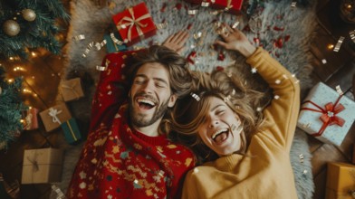 Young couple laying on their backs surrounded by christmas gifts and decorations laughing.