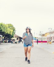 Smiling young tourist in hat walking with cell phone on the street. Female tourist texting phone in