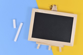 Empty wooden black chalkboard next to pieces of white chalk on yellow and blue background