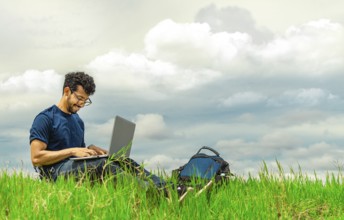 Smiling freelancer man sitting with laptop and backpack in field. Portrait of a man sitting on the