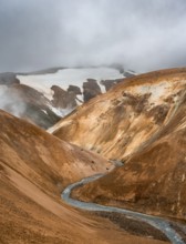 Steaming stream between colourful rhyolite mountains with snowfields, Hveradalir geothermal area,