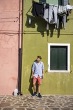 Young man leaning against a house wall, green and pink house facade with laundry to dry, colourful