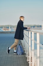 Young student with backpack in old wool coat, scarf and short trousers leaning to the railings