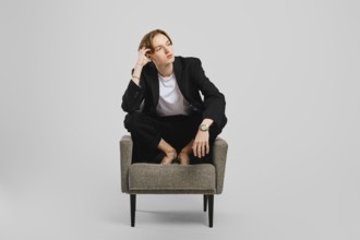 Young barefoot man in black suit sitting in armchair in studio