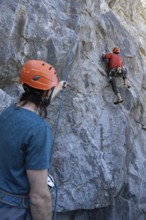 Young man tries to climb a rock while his colleague holds the safety harness