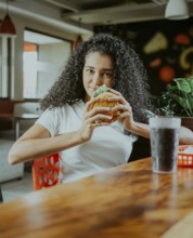 Beautiful latin girl enjoying a delicious hamburger in a restaurant. Portrait of afro girl holding