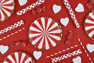 Party flat lay with paper plates, heart ornaments, candy and drinking straws on red background