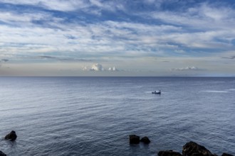 A small boat sails on a calm sea under a blue sky with few clouds, surrounded by stones, fishing