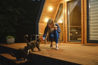 Cute woman sits on a wooden deck and playing with two curious cats