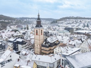 Snow-covered town view with central church tower and surrounding buildings in a hilly environment,