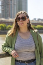 Portrait of a young woman wearing large sunglasses, a striped tank top, and a green jacket,