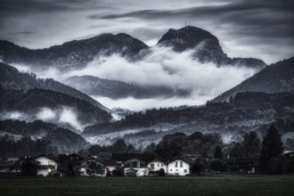 A dramatic landscape with mist-covered mountains, forests and a small village in the foreground,