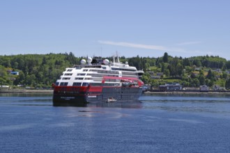 A large Hurtigruten ship sails along a forested coastline under a clear sky, Cormorant Island,