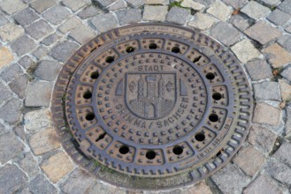 Round manhole cover with city coat of arms, surrounded by cobblestones, manhole cover, manhole