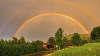 A double rainbow spans a green landscape with trees and houses at dusk, Bad Feilnbach