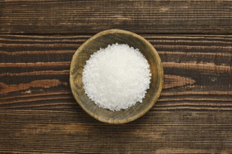 Coarse salt is displayed in a rustic wooden bowl, set against a textured wooden table. The natural