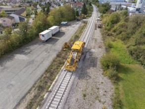 A yellow construction machine on a railway track in a residential area with some lorries nearby,