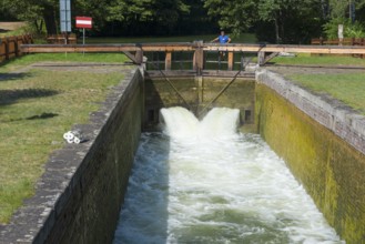 Strong water flow through an open lock with a wooden bridge, surrounded by a green park, Sluza