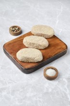 Three breaded cutlets on a rustic wooden cutting board, accompanied by small bowls of salt and