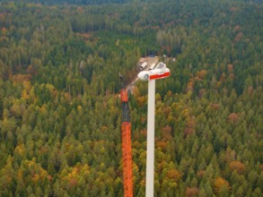 A crane assembles the upper part of a wind turbine above an autumnal forest, wind farm construction