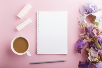 Purple and blue iris flowers and a cup of coffee with notebook on pastel pink background. Morninig,