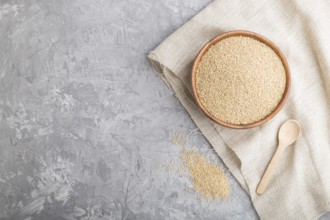 Wooden bowl with raw white quinoa seeds and wooden spoon on a gray concrete background and linen