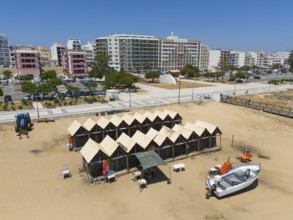 Beach with fishing huts and fishing boats, residential buildings and a clear blue sky behind,