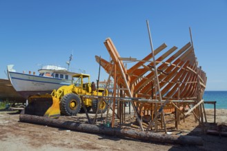 A ship under construction with a yellow excavator at a shipyard by the sea under a blue sky,