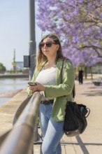 A vertical shot of a woman wearing sunglasses and a green jacket, holding her smartphone with both