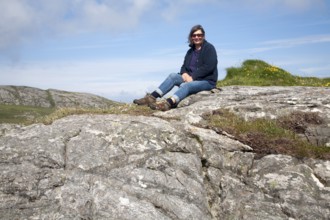 Middle aged female hiker sitting high on rocky outcrop in Barra, Outer Hebrides, Scotland, UK